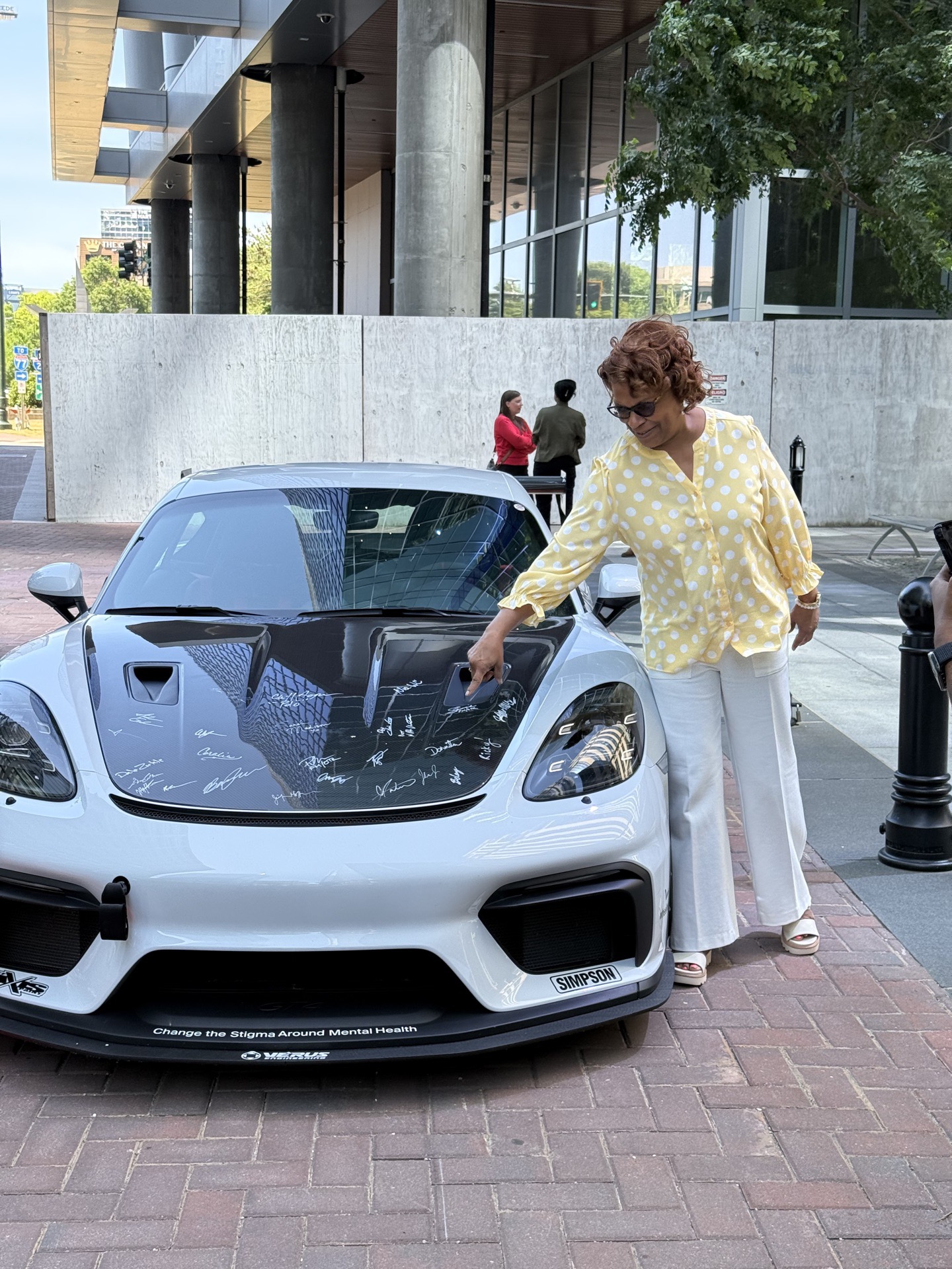 Attendee in a yellow polka-dot dress signs the Team Wheelhouse signed-hood Cayman GT4 on the green at Legacy Union
